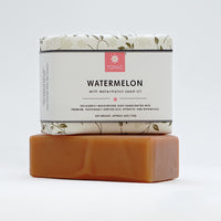 Watermelon bar soap wrapped in floral paper, stacked on top of a unwrapped bar of soap, against a white background.